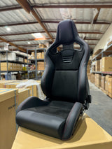 A black racing bucket seat with red contrast stitching, displayed on a cardboard box in a warehouse setting.